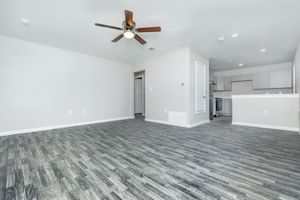 Spacious, well-lit living area featuring a ceiling fan, light hardwood flooring, and neutral-colored walls. An open layout connects to a modern kitchen with white cabinetry. A doorway leads to another room, enhancing the sense of space.
