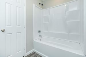 A clean and modern bathroom featuring a white bathtub with a shower combination. The walls are a light color, and there is a single door leading to an adjacent area. The floor is covered with wood-like tiles, adding a touch of warmth to the space.