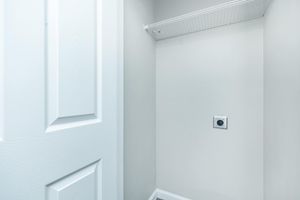 An empty closet with light gray walls, a white door, and a wire shelf mounted on the wall. There is a power outlet visible on the wall, indicating potential use for appliances. The floor is not captured in the image, creating a clean and minimalistic appearance.
