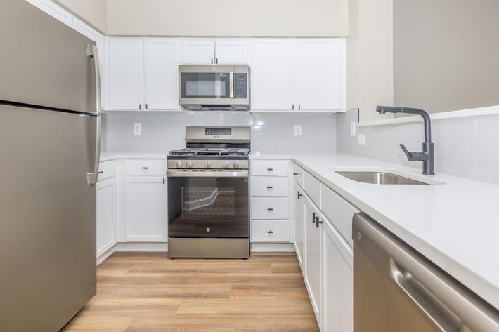 Modern kitchen featuring white cabinetry, stainless steel appliances including a refrigerator, microwave, and oven, along with a dishwasher. The countertop is light-colored and smooth, complemented by a sleek kitchen faucet. The floor is finished with wooden planks, creating a warm atmosphere.