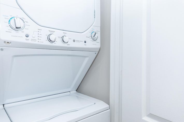 A stacked washer and dryer unit in a laundry room, with control knobs for different settings. The appliance is white and positioned next to a closed door. The overall setting is clean and modern.