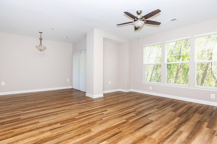 A spacious room featuring a light-colored wall, large windows with blinds, and a ceiling fan. The floor is made of polished wood, giving the space a warm appearance. There is a light fixture hanging from the ceiling, and a sliding door is partially visible on the left side, leading to a potential outdoor area.