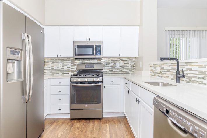Modern kitchen featuring stainless steel appliances including a refrigerator, microwave, and gas stove. The cabinetry is white, complemented by a stylish backsplash with mosaic tiles. A sleek sink with a modern faucet sits on a light-colored countertop, and the floor is a warm wood finish. Natural light enters through a window.