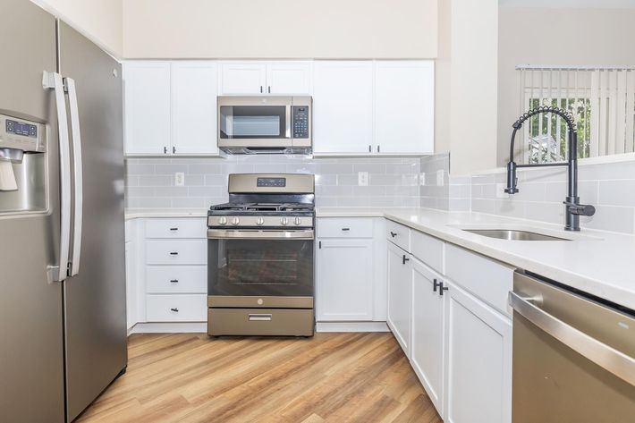Modern kitchen with stainless steel appliances, including a refrigerator, stove, and microwave. White cabinetry complements the sleek design, while gray tiled backsplash adds a stylish touch. The countertop is spacious, and the kitchen features warm wood flooring for an inviting atmosphere. Bright and airy with plenty of natural light.