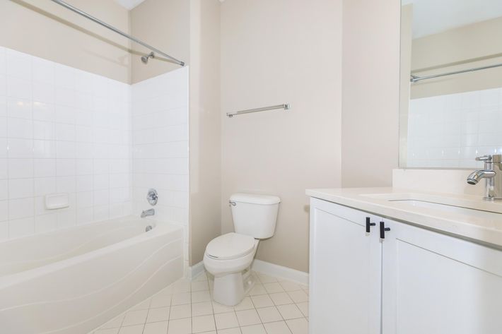 A clean and modern bathroom featuring a bathtub with a shower, a white toilet, and a sink vanity with a mirror. The walls are painted in a light beige color, and the floor is tiled in a white pattern. A towel rack is mounted on the wall beside the bathtub.