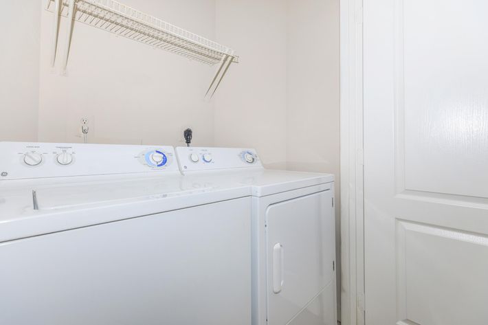 A laundry room featuring a white washing machine and dryer side by side. Both appliances have control dials and are set against a wall with a small shelf above for storage. A white door is visible in the background.
