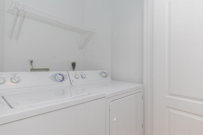 A clean, well-lit laundry room featuring a white washing machine and dryer side by side. Above them, a wire shelf provides additional storage space. The walls are painted a light color, and a door is partially visible, indicating a neat and organized environment.