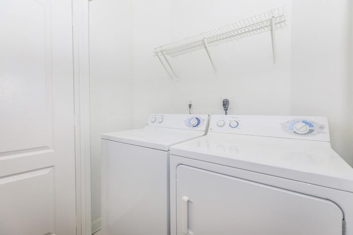 A clean and modern laundry room featuring a white washer and dryer side by side. Above them, there is a wire shelf for storage. The walls are painted white, creating a bright and tidy atmosphere, with a closed door visible on the left side.