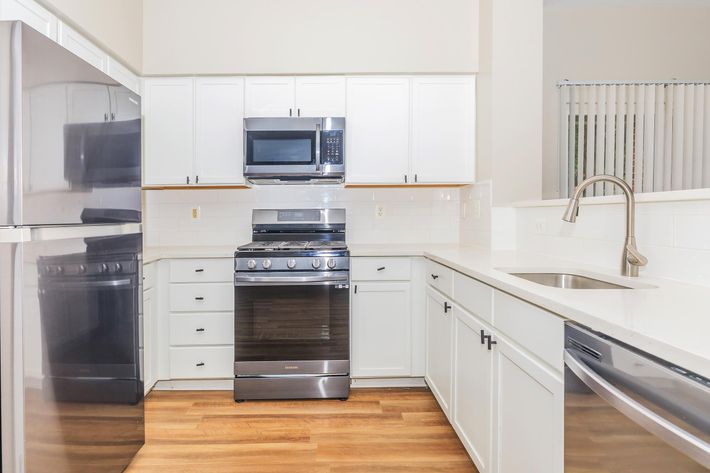Modern kitchen featuring white cabinetry, stainless steel appliances including a microwave and an oven, and a spacious countertop. The floor is made of light wood, and there is a sink with a sleek faucet. Natural light enters through a window with blinds.