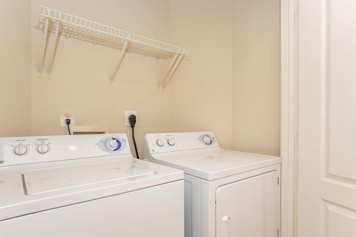 A laundry room featuring a white washing machine and a white dryer side by side. Above them is a wire shelf for storage. The walls are painted a neutral color, and a door is visible in the background. The space is clean and organized, suitable for household laundry tasks.