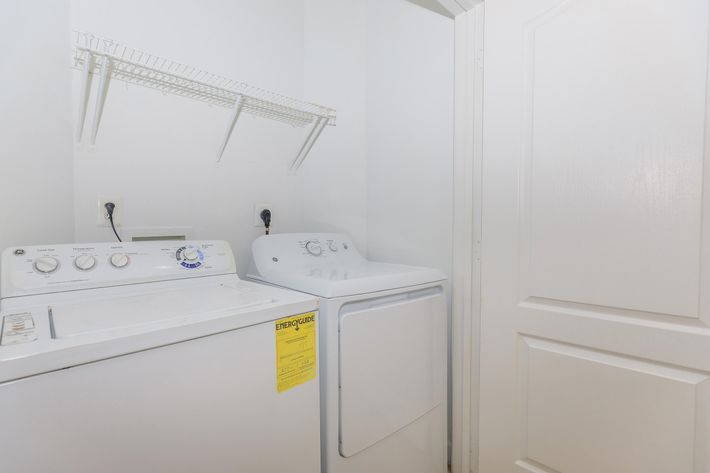 A small laundry area featuring a washer and dryer side by side. The washer is on the left with knobs and a switch panel visible, while the dryer is on the right, labeled with a tag. Above, there is a wire shelf attached to the wall. The walls are painted white, enhancing the bright and clean appearance of the space.