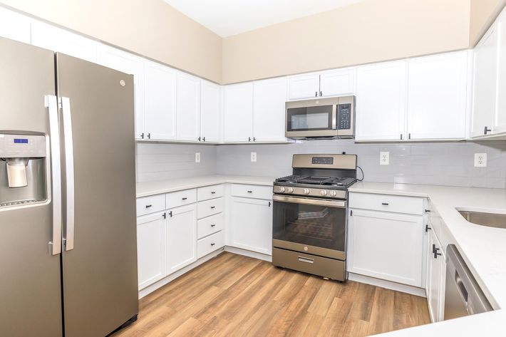 Modern kitchen featuring white cabinets, a stainless steel refrigerator with a water dispenser, a gas stove, and a microwave oven. The countertops are light-colored with a wooden floor, creating a bright and inviting space. The walls are painted a neutral shade for a contemporary look.