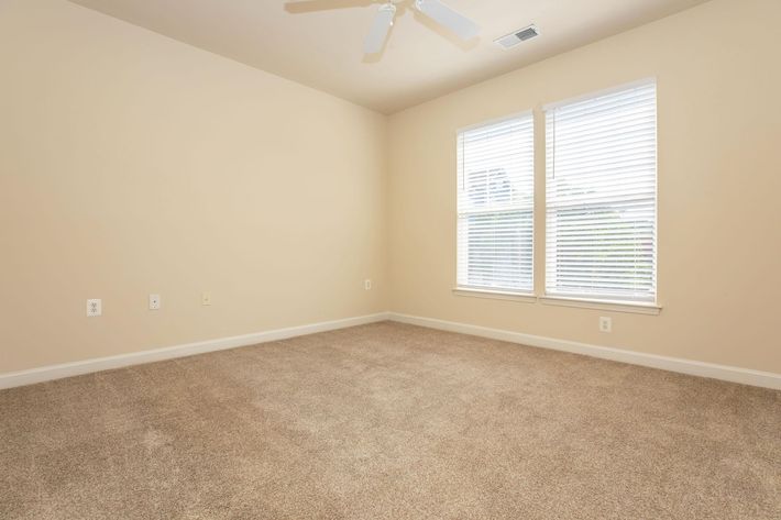 A well-lit, empty room with beige walls and light-colored carpet. Two windows with white blinds allow natural light to enter. The ceiling fan is visible, and there are no furnishings in the space, creating a minimalist and open atmosphere.