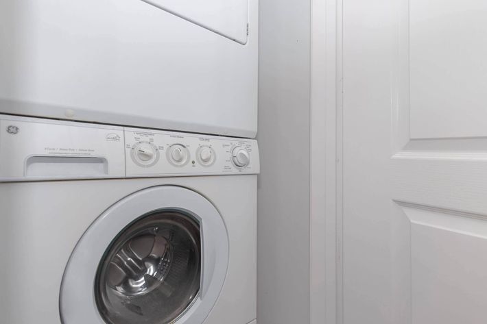 A stacked washer and dryer in a laundry room, featuring a white exterior and control knobs for settings. The appliances are positioned next to a closed white door, with a neutral-colored wall in the background.
