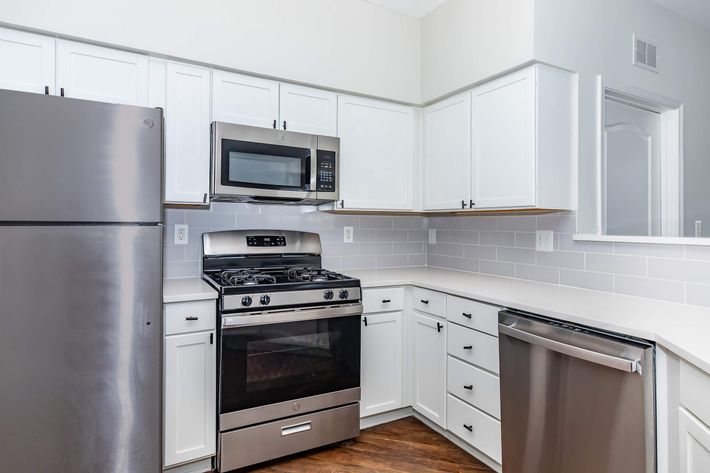 Modern kitchen featuring stainless steel appliances, including a refrigerator, microwave, and gas stove. The cabinets are white, with a sleek countertop and backsplash of light tiles. The floor is a warm wood finish, creating a bright and inviting space.