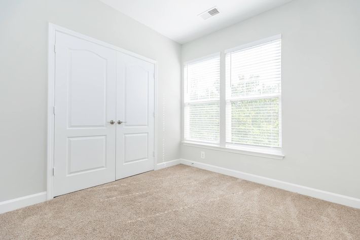 A bright, empty room with light gray walls and a tan carpet. There are two large windows with white blinds allowing natural light to fill the space. A double door closet is visible on one wall, adding to the room's simplicity and openness.
