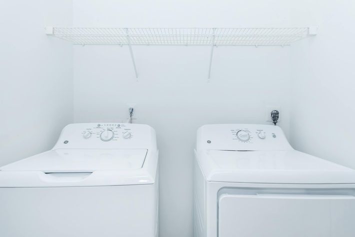 A clean and modern laundry room featuring a white washing machine and a white dryer side by side. Above them, a simple wire shelf is mounted on the wall for storage. The walls are painted white, creating a bright and spacious atmosphere.