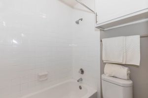 A clean, modern bathroom featuring a white tiled shower area with a chrome showerhead, a bathtub with a silver faucet, and a toilet. Soft white towels are neatly stacked on a shelf above the toilet, enhancing the overall bright and minimalistic design.