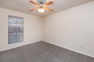 Empty room with light-colored walls and a ceiling fan with wooden blades. A window with blinds allows natural light to enter, and the floor is covered in gray carpeting. The room has no furniture or decorations, creating a spacious and minimalistic appearance.