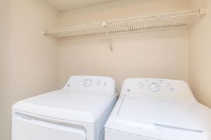 A clean laundry room featuring a stacked washer and dryer set in white. Above them is a wire shelf for storing laundry supplies. The walls are a neutral color, creating a bright and simple space.