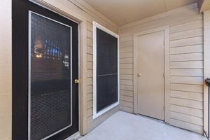 A view of a small porch area featuring two doors—one with a black frame and a screen and another in a beige color. The walls are wooden with a light brown finish, and the floor is concrete. Natural light is visible, suggesting a welcoming entrance.