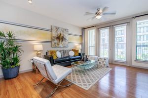 A modern living room featuring a black sofa with yellow and gray cushions, a glass coffee table on a patterned rug, and stylish chairs. Natural light floods in through large windows, enhancing the warm wood flooring and decorative wall art. A tall plant adds a touch of greenery to the space.
