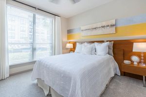 A cozy bedroom featuring a queen-sized bed with white bedding and several decorative pillows. The walls are painted in soft tones with a horizontal artwork above the bed. Two bedside tables with lamps flank the bed, and large windows with blinds let in natural light, creating a bright and inviting atmosphere.