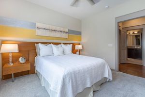 A cozy bedroom featuring a queen-sized bed with a white bedding set, flanked by two bedside tables with lamps. The wall behind the bed has a decorative painting with horizontal stripes in gray and yellow. A door leads to a bathroom, and the room has neutral-colored carpet and walls.