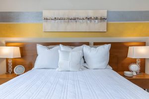 A neatly made bed with fluffy white pillows and a textured white blanket. On either side of the bed, there are stylish lamps with soft light. Above the headboard, a modern artwork in neutral tones adds a decorative touch to the room, while a small clock and decorative items sit on the nightstands.