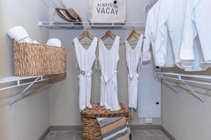 A well-organized closet featuring three white dresses hanging neatly on hangers, a woven basket holding towels or linens, a stylish hat on a shelf above, and a sign that reads "ALWAYS ON VACAY". A few white shirts are also hanging, creating a fresh, inviting atmosphere.