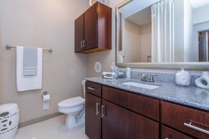 Modern bathroom featuring a dark wooden vanity with a granite countertop. There's a round mirror above the sink and a light-colored shower curtain. A white towel hangs neatly on a rack, and a toilet is positioned next to the vanity. A decorative container holds cotton balls on the counter. Spacious and well-lit.
