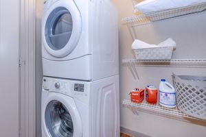 A stackable washer and dryer unit in a laundry area, surrounded by shelves containing cleaning supplies, including laundry detergent and bleach. There's a basket and a white container on the shelves, with rolls of towels stacked nearby. The walls are plain, and the floor is wooden.