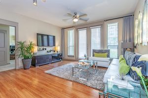 A stylish living room featuring a light gray sofa with decorative pillows, a glass coffee table on a plush rug, and a sleek black TV cabinet. Large windows adorned with sheer curtains allow natural light to fill the space, complemented by potted plants and modern art on the walls. The flooring is warm wood, enhancing the cozy atmosphere.