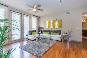 A modern living room featuring a white sectional sofa with green and gray cushions, a glass coffee table on a plush gray rug, and large windows with blinds. Decorative plants and contemporary wall art enhance the cozy atmosphere, while a ceiling fan adds a touch of functionality.