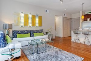 A modern living room featuring a white sofa with decorative pillows, a glass coffee table, and a plush area rug. The walls are adorned with abstract art, and there are stylish lamps on side tables. A kitchen area with bar stools is visible in the background, enhancing the open-concept design.