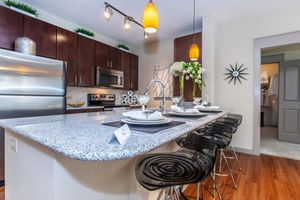 Modern kitchen featuring dark wood cabinetry and stainless steel appliances. A granite countertop with four black bar stools is set for a meal. Decorative items, including a vase with flowers and stylish lighting fixtures, enhance the space's contemporary feel.