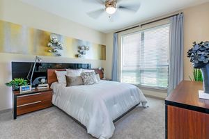 A modern bedroom featuring a queen-sized bed with white bedding, decorative pillows, and a wooden headboard. Dark wood nightstands flank the bed, and a stylish lamp sits on one. The room has a large window with blinds, allowing natural light to enter, and includes decorative wall art and green plants for a fresh touch.