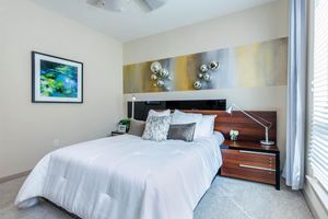 A modern bedroom featuring a neatly made bed with a white bedspread, a wooden headboard, and stylish bedside tables. The decor includes a colorful abstract painting above the bed and decorative round wall pieces. Natural light streams in through a window with blinds, enhancing the inviting atmosphere.