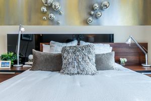 A modern bedroom featuring a neatly made bed with textured pillows, a wooden headboard, and stylish bedside lamps. Behind the bed, decorative metallic spheres are mounted on a gold-accented wall. A small plant and a framed photo are placed on the nightstand, adding a touch of decor.