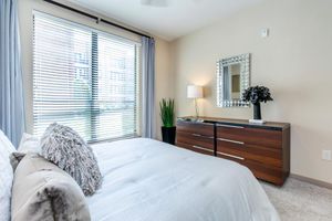 A softly lit modern bedroom featuring a neatly made bed with decorative pillows, a sleek wooden dresser, a minimalist lamp, and a decorative plant. Large windows with blinds provide natural light, enhancing the serene atmosphere, while a framed mirror adds an elegant touch to the decor.
