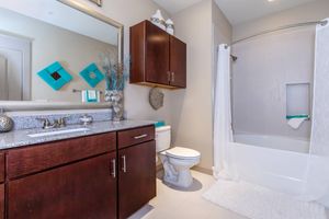 A modern bathroom featuring a granite countertop with a sink, wooden cabinets, a toilet, and a shower with a white curtain. The walls are light-colored, and there are decorative teal accents, including wall art and towels. The floor is tiled, and a soft white rug lies near the bathtub.