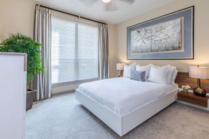 A well-decorated bedroom featuring a white platform bed with layered pillows, two bedside lamps, and a stylish piece of wall art. A potted plant adds greenery, and large windows with blinds allow natural light to filter in, enhancing the cozy ambiance with soft, neutral-colored decor.