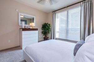 A modern bedroom featuring a queen-sized bed with white linens, a decorative gray pillow, a dresser with a wooden top, and a potted plant. Large windows with blinds allow natural light in, complemented by light-colored walls and a ceiling fan, creating a bright and inviting atmosphere.