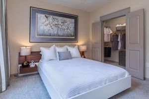 A cozy bedroom featuring a white bed with plush pillows, a decorative wall art piece depicting blossoms, and bedside lamps. A spacious closet with organized clothing is visible through an open door, and the room is decorated in neutral tones with soft, carpeted flooring.