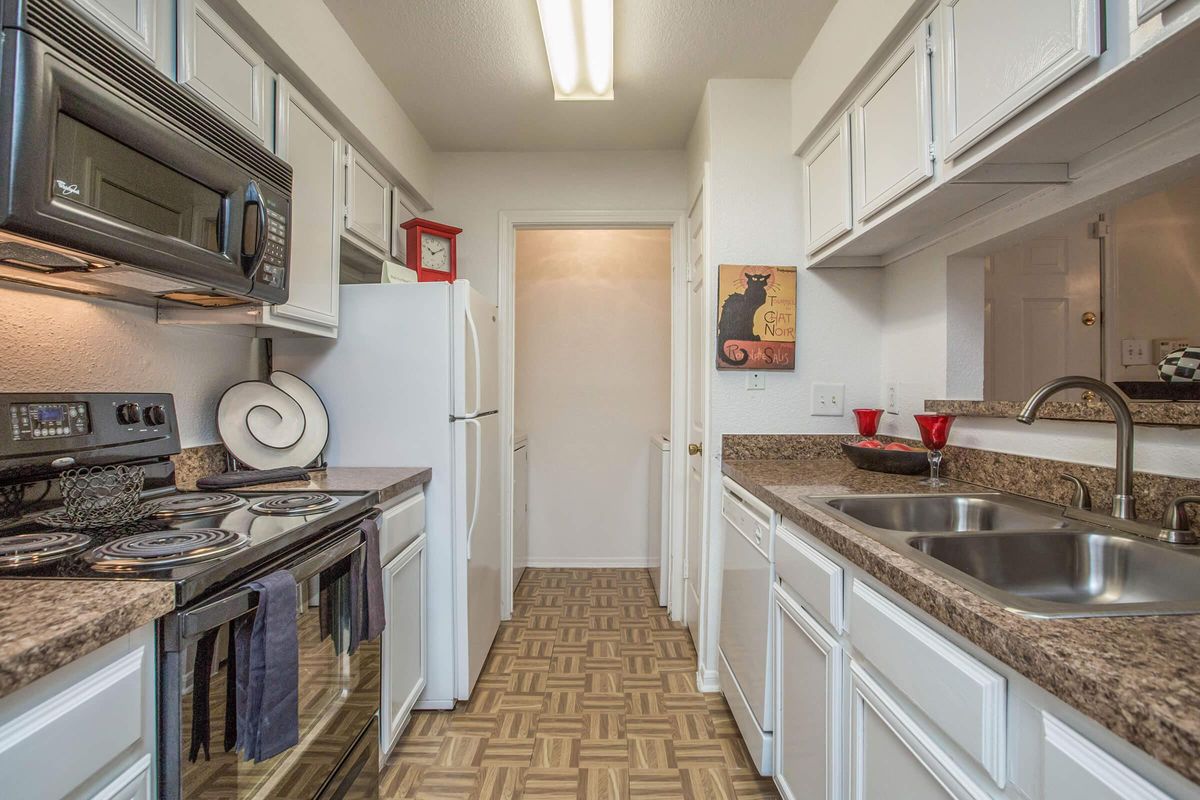 A modern kitchen featuring white cabinetry, a dark countertop, and stainless steel appliances. It includes a stove, microwave, and refrigerator, with decorative items like a bowl and red accessories on the counter. The floor has a geometric pattern, and a doorway leads to another room.