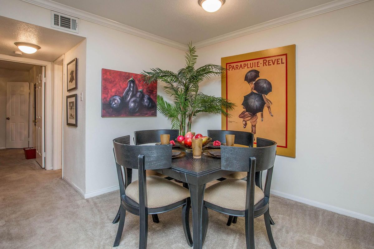 A dining area featuring a round table with four chairs, adorned with a centerpiece of fruit in bowls. The walls display a colorful painting of fruit and another artwork featuring umbrellas. A potted plant adds greenery to the space, which is softly lit and has a neutral wall color, enhancing the inviting atmosphere.