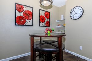 A cozy dining nook featuring a dark wood round table with black leather stools. Two framed red flower wall art pieces hang on the light-colored wall, alongside a wall clock. A small shelf displays decorative items, and a bowl of colorful fruit is centerpiece on the table, adding a warm, inviting touch.