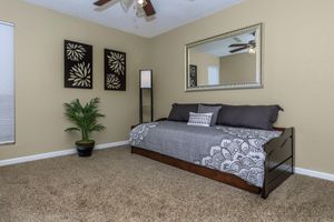 A cozy bedroom featuring a daybed decorated with gray and white bedding. On the wall, there are two floral pattern wall hangings and a large mirror. A tall floor lamp stands beside the bed, and a potted plant adds a touch of greenery. The room is painted in a warm beige tone, with neutral carpet flooring.