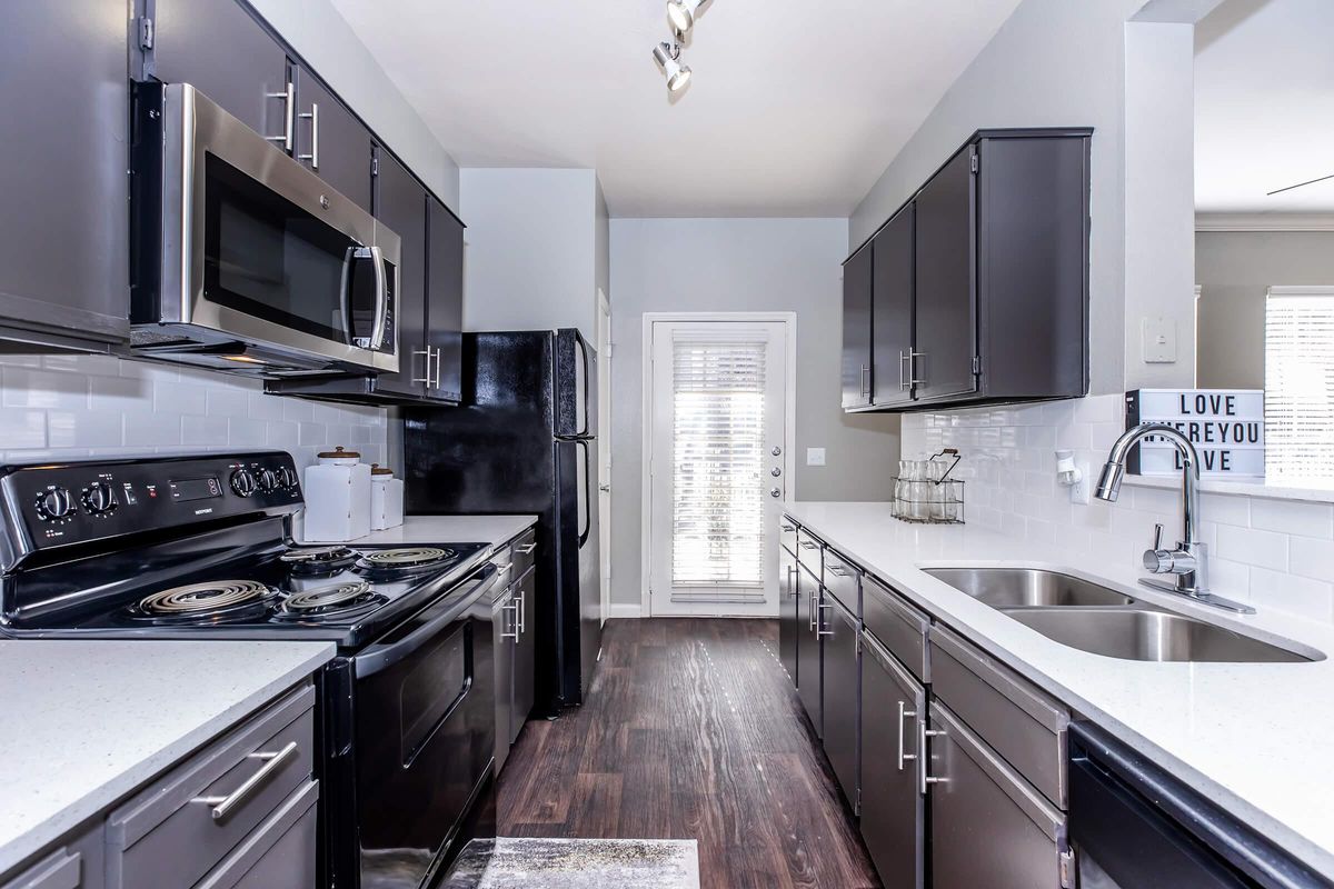 Modern kitchen featuring dark cabinets, stainless steel appliances, and light countertops. Includes a black stove, microwave, and refrigerator. The space has a window with blinds, allowing natural light, and a decorative wall sign that says "LOVE" in white letters. The floor is dark wood laminate.