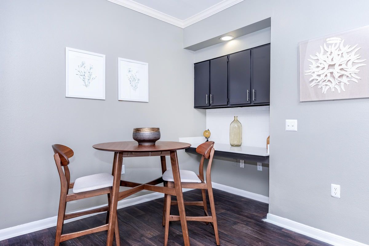 A modern dining area featuring a round wooden table with two high-backed chairs. There are decorative items on the table and a light-colored vase in the background. The walls are painted a soft gray, and there are framed botanical prints on the wall. The flooring is dark wood, adding warmth to the space.